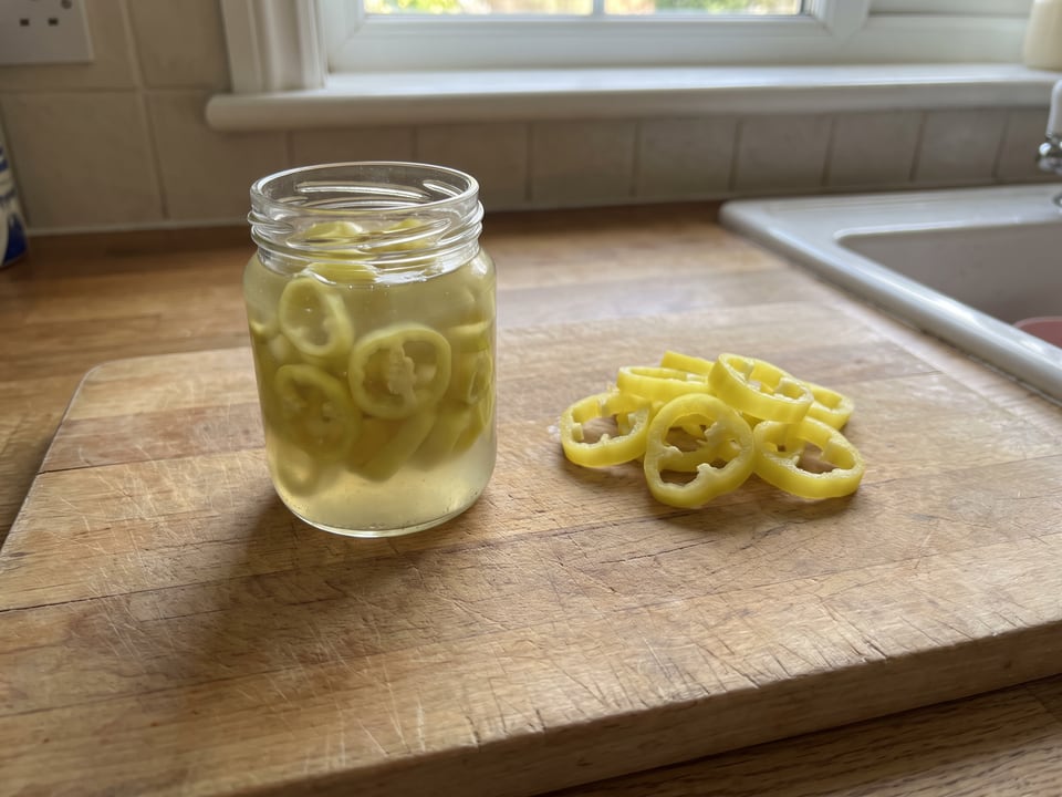 A jar of brined pale pepper rings next to a pile of loose fresh yellow pepper rings